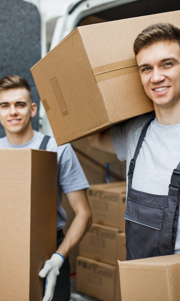 Two young handsome smiling workers wearing uniforms are standing in front of the van full of boxes
